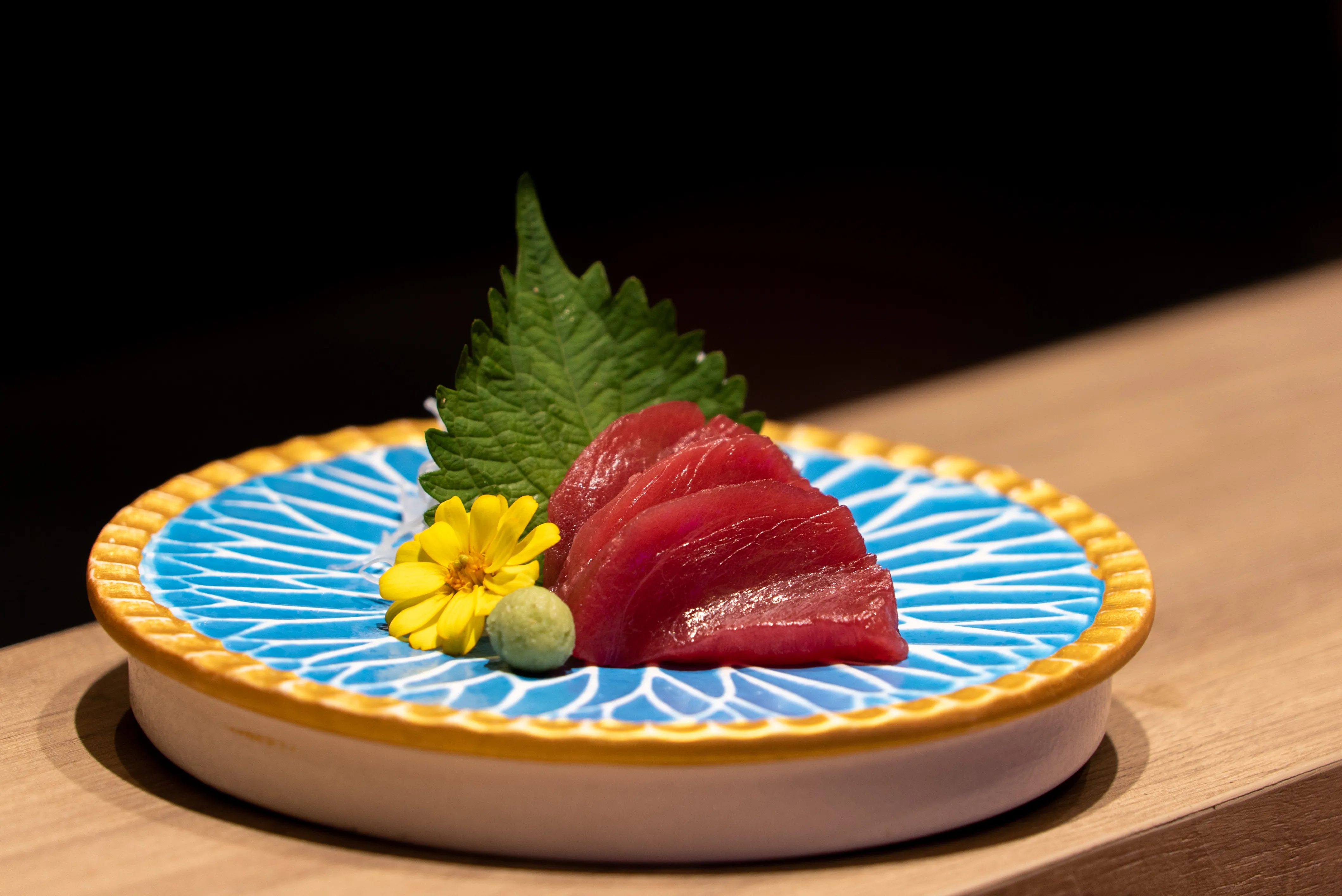 Four slices of deep red akami tuna sashimi are elegantly arranged on a vibrant blue and gold patterned plate, garnished with a green shiso leaf, shredded daikon, a yellow edible flower, and a dollop of fresh green wasabi.