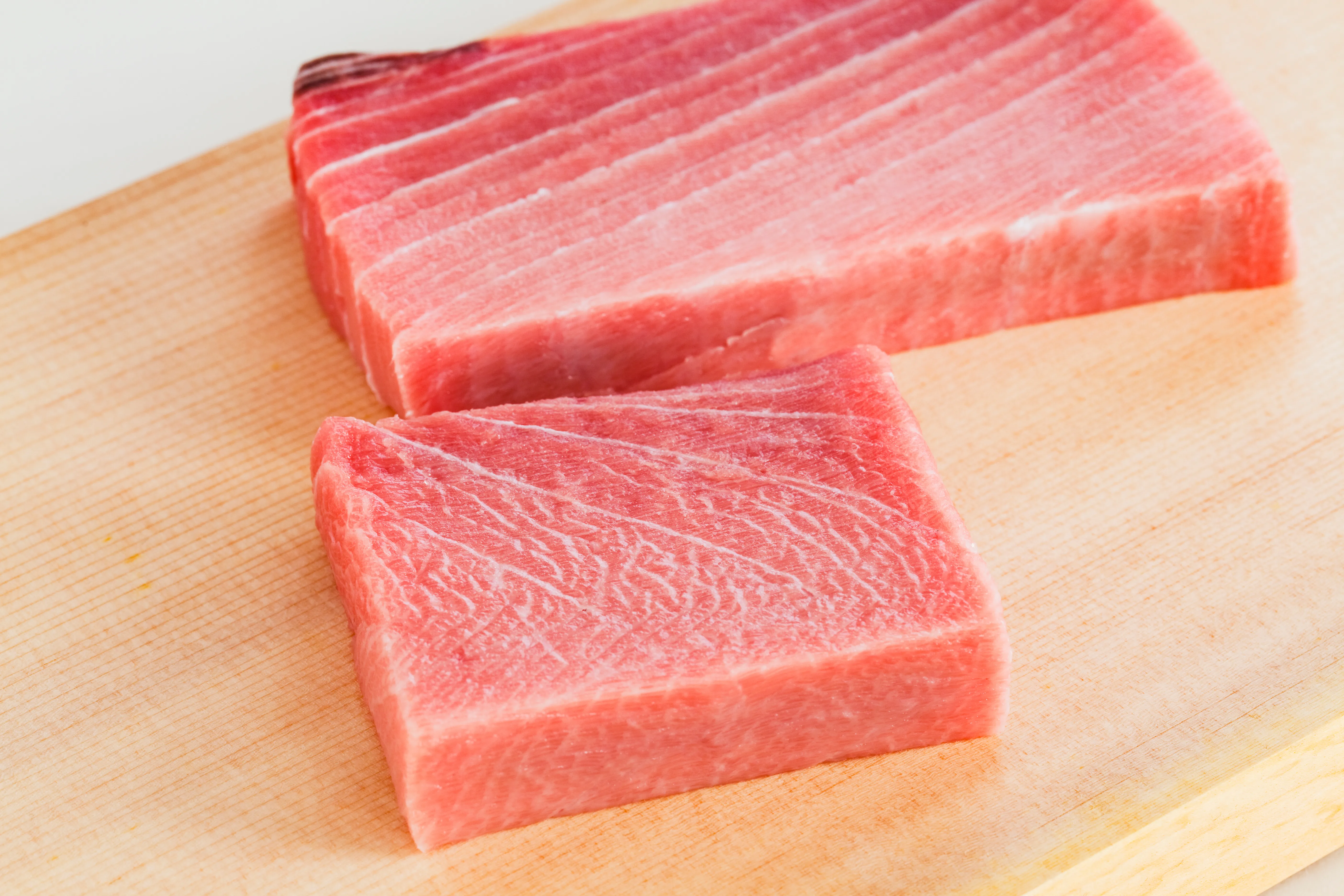 Two rectangular blocks of fresh, moderately marbled Chutoro tuna lie on a light wooden cutting board, ready for preparation.