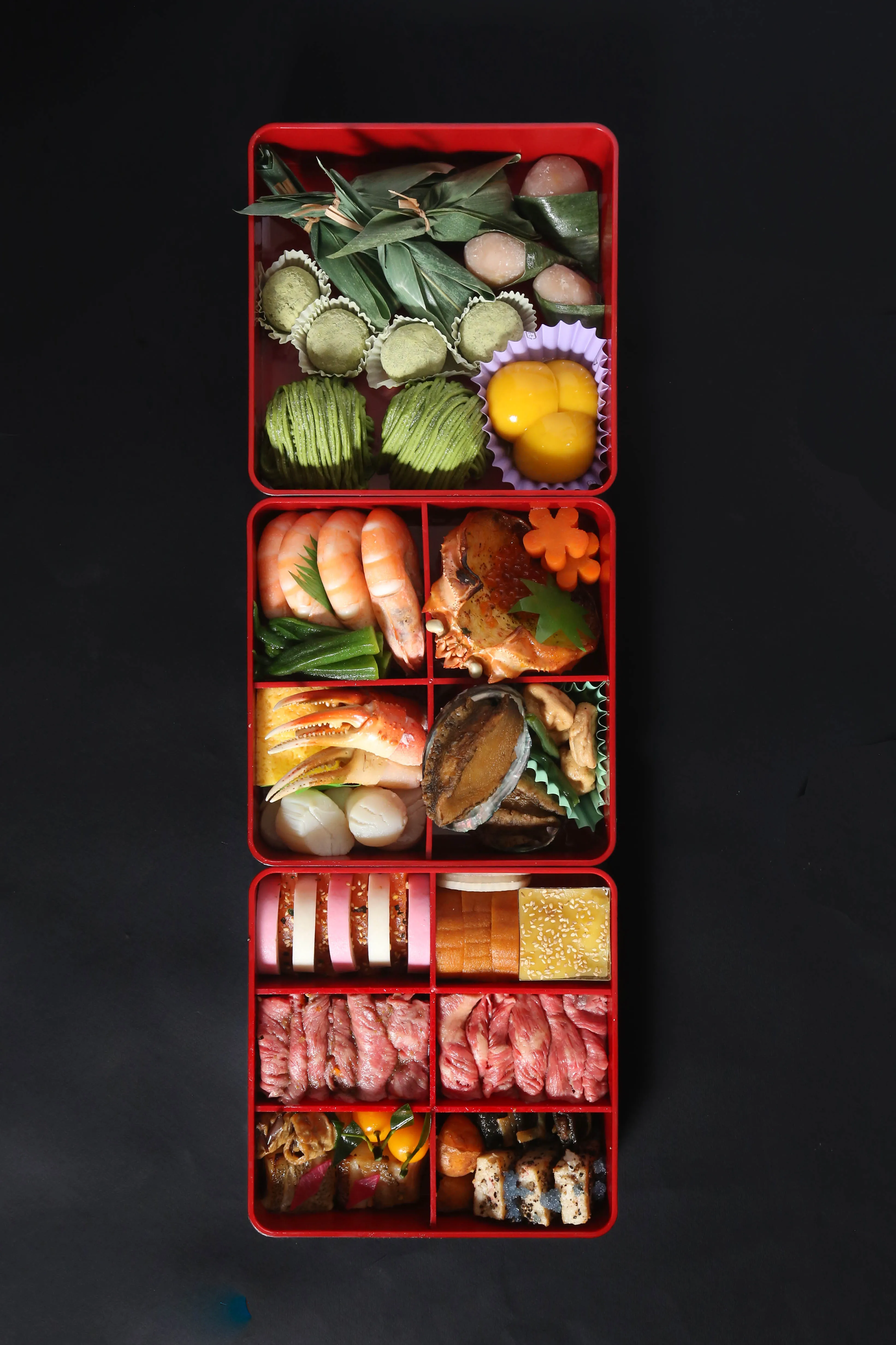 A vibrant, multi-tiered Osechi Ryori box on a black background, filled with traditional Japanese New Year foods.