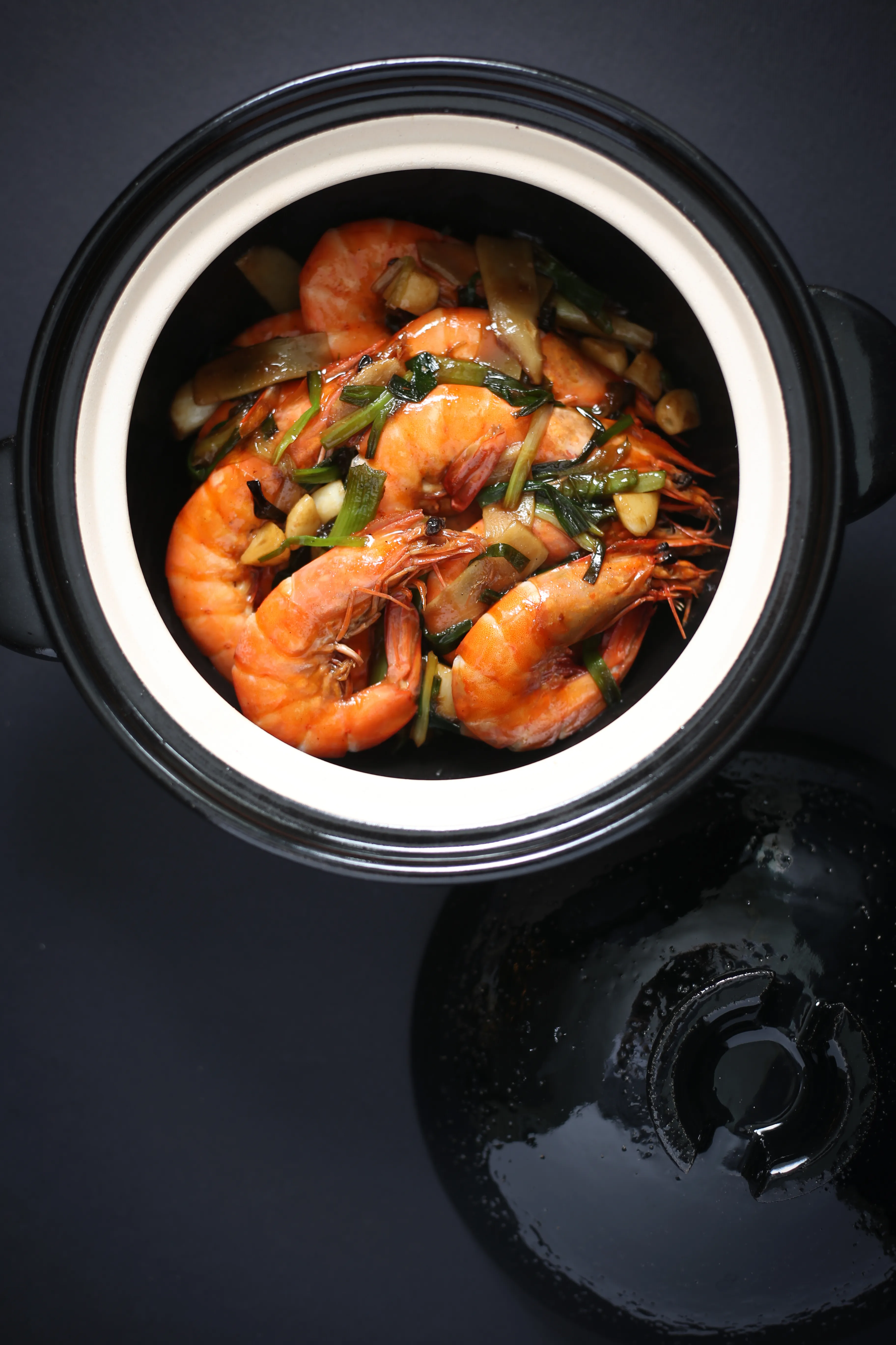 An overhead shot of vibrant orange shrimp, fresh green scallions, and sliced ginger stir-fried in a black ceramic claypot with a creamy white rim, set against a dark background.