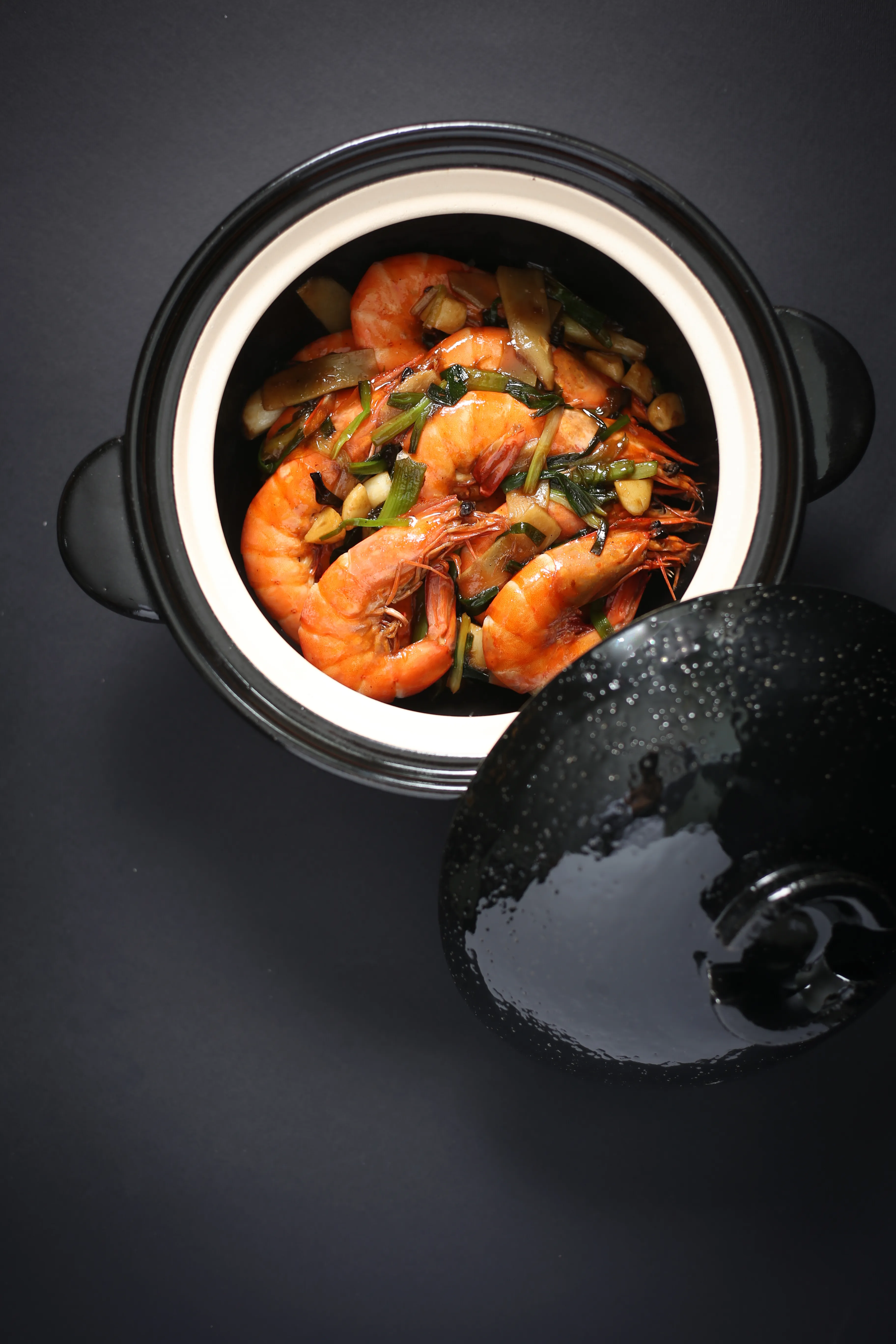An overhead shot of a black ceramic pot filled with plump, vibrant orange shrimp, green scallions, and slices of ginger in a rich, glossy sauce. The pot's textured black lid rests next to it on a dark, minimalist background.