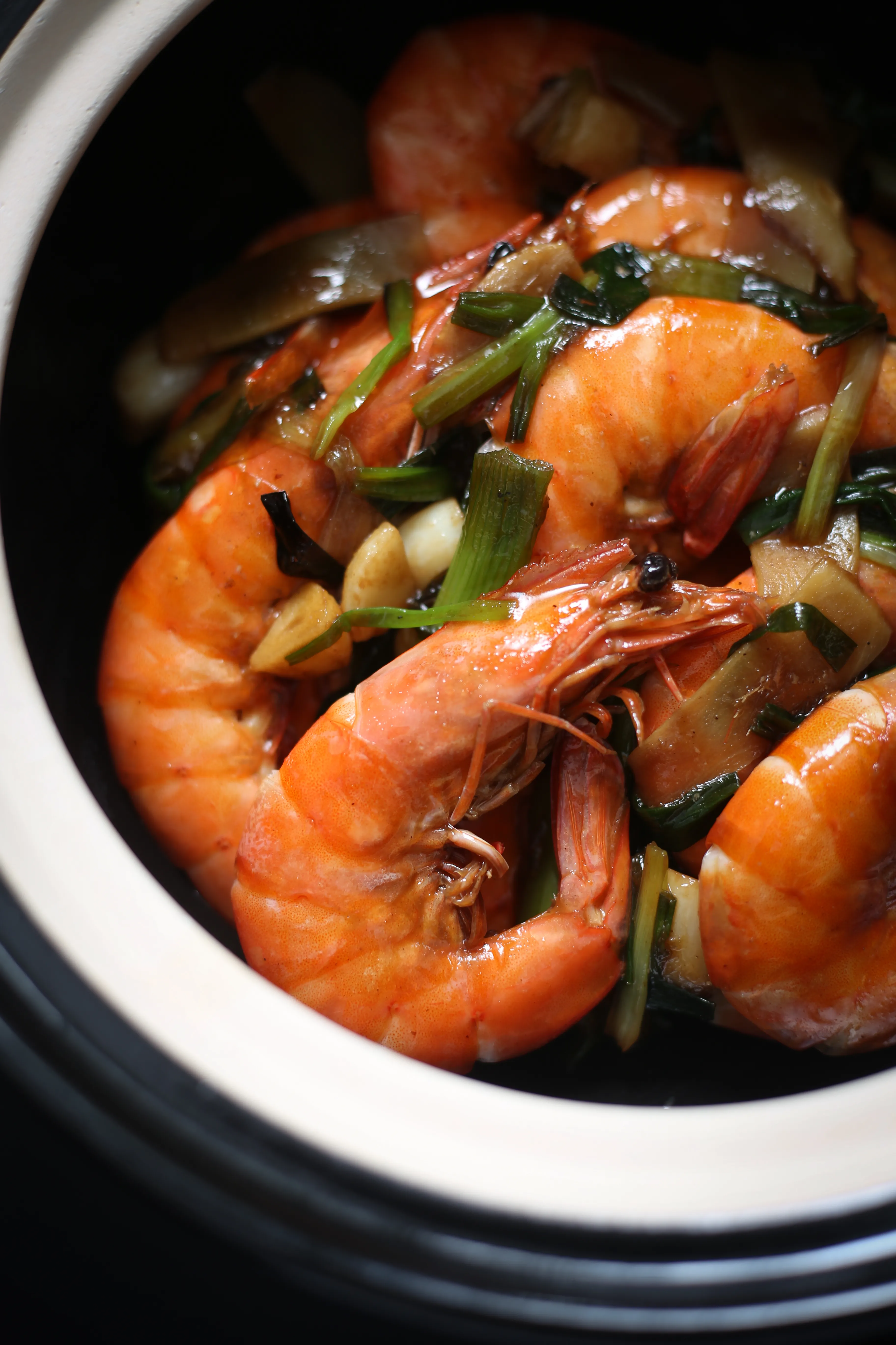 An overhead shot of plump, cooked shrimp with sliced ginger and green scallions, served in a sleek black ceramic pot with a cream-colored rim, set against a dark background.