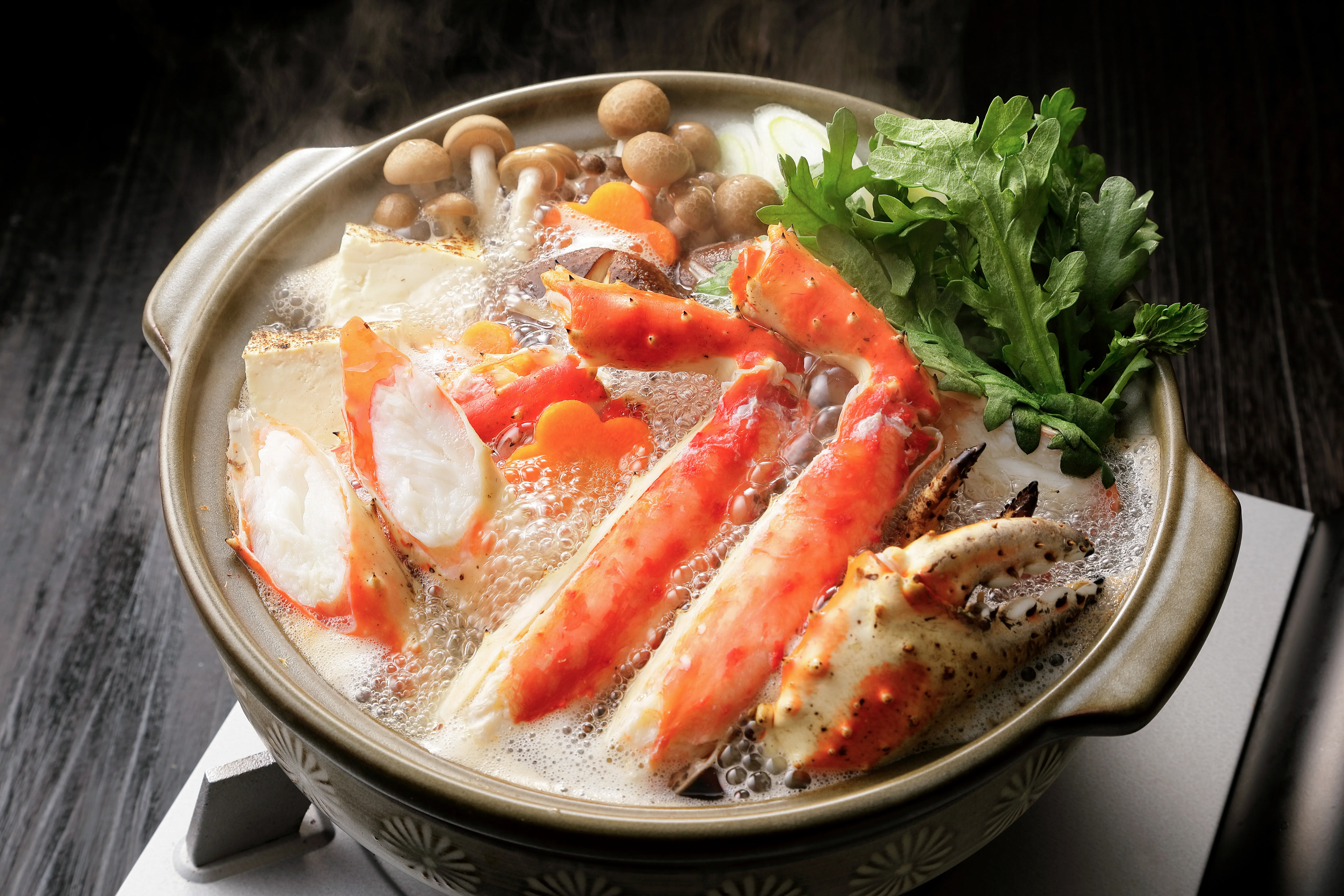 A bubbling Japanese hotpot filled with vibrant red king crab legs, fresh green vegetables, mushrooms, and tofu, emitting steam against a dark background.