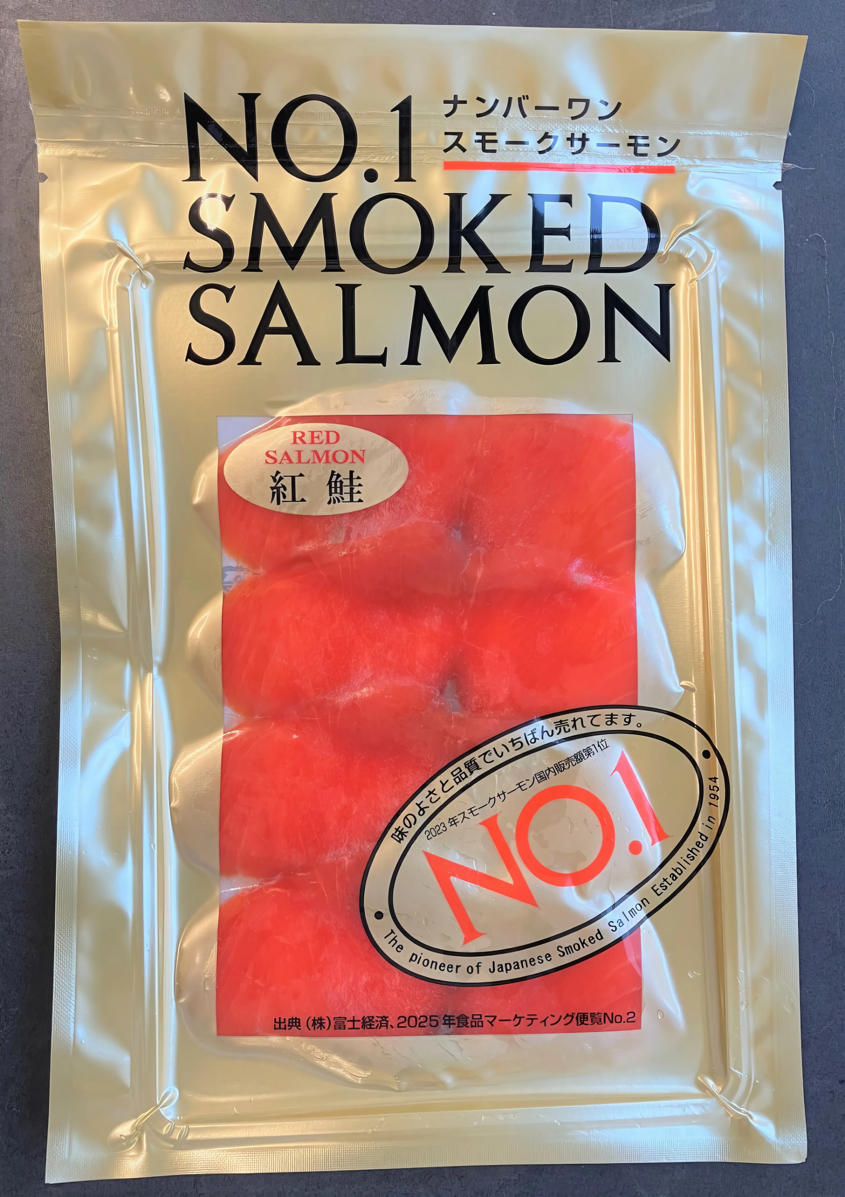 A gold-colored vacuum-sealed package of 'NO.1 Smoked Salmon' from Japan, with five vibrant red salmon fillets visible through a clear window. The packaging features Japanese text and states 'The pioneer of Japanese Smoked Salmon Established in 1954'.