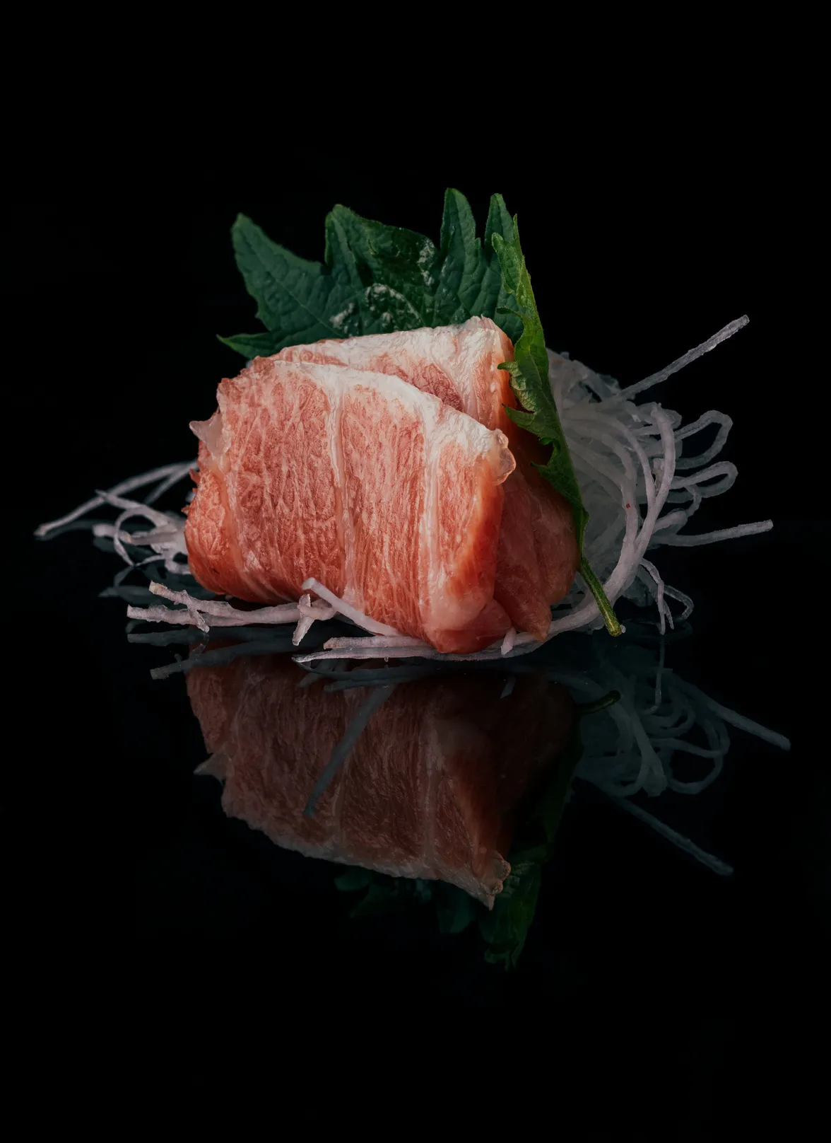 Two thick slices of premium otoro tuna sashimi are elegantly presented on a bed of finely shredded daikon radish with a vibrant green shiso leaf, against a reflective black background.