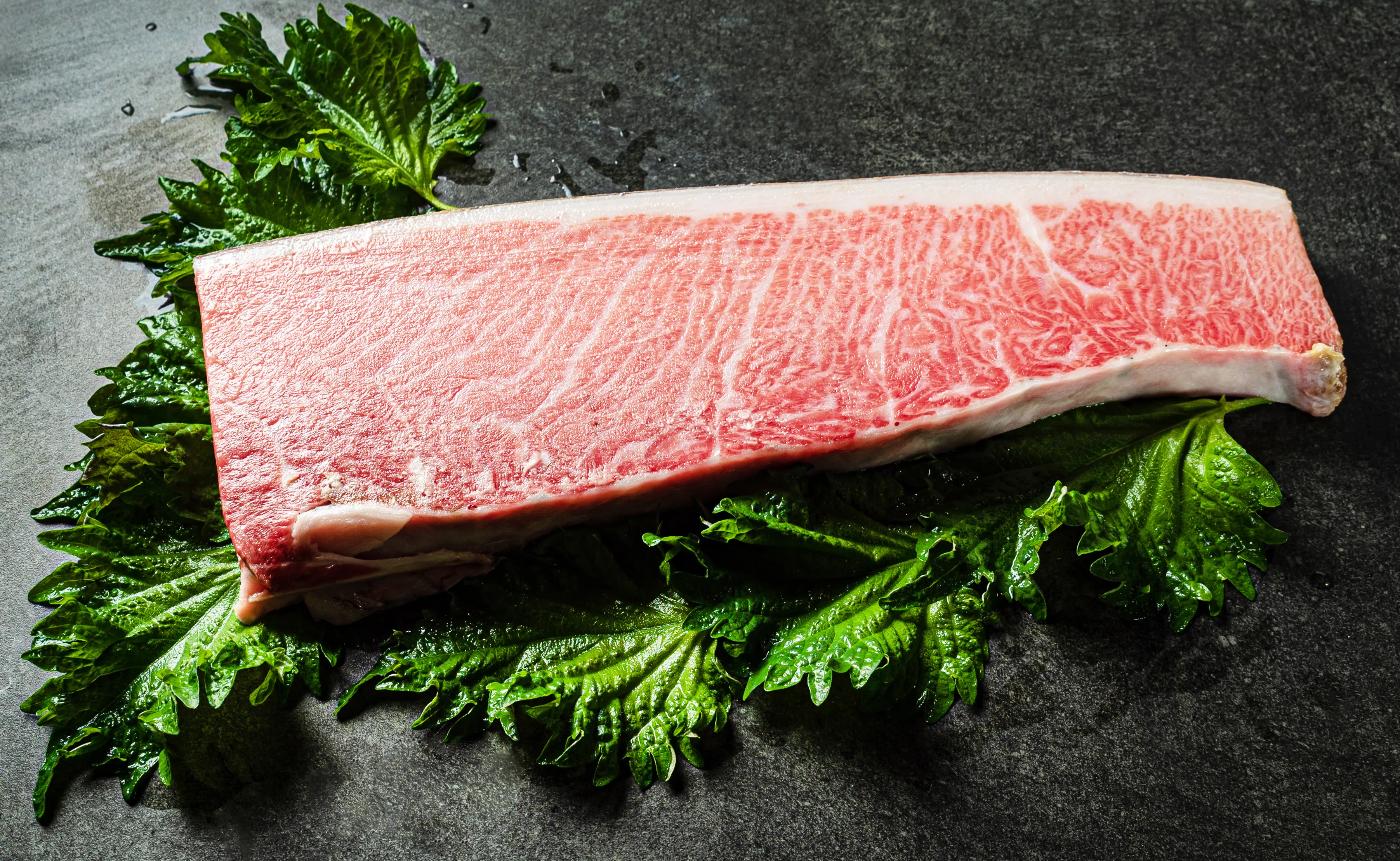 A large, rectangular block of intensely marbled pink and white bluefin otoro tuna rests on vibrant green shiso leaves, atop a dark, textured stone surface with scattered water droplets.