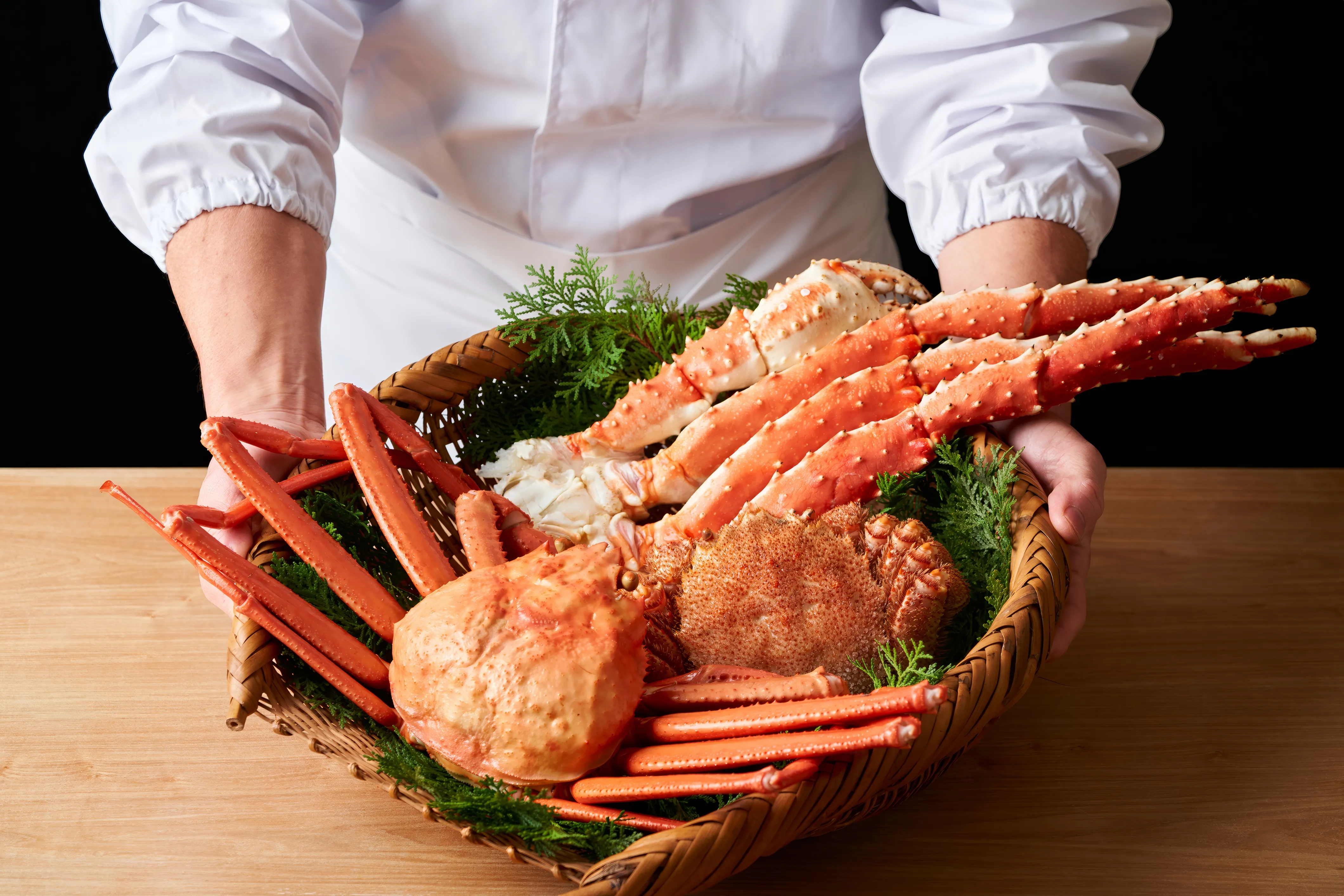 A chef in a clean white uniform holds a woven bamboo basket overflowing with vibrant red and orange premium Japanese crab, including large King Crab legs and a whole Hairy Crab, garnished with dark green foliage. The scene is set on a light wooden surface against a dark background.