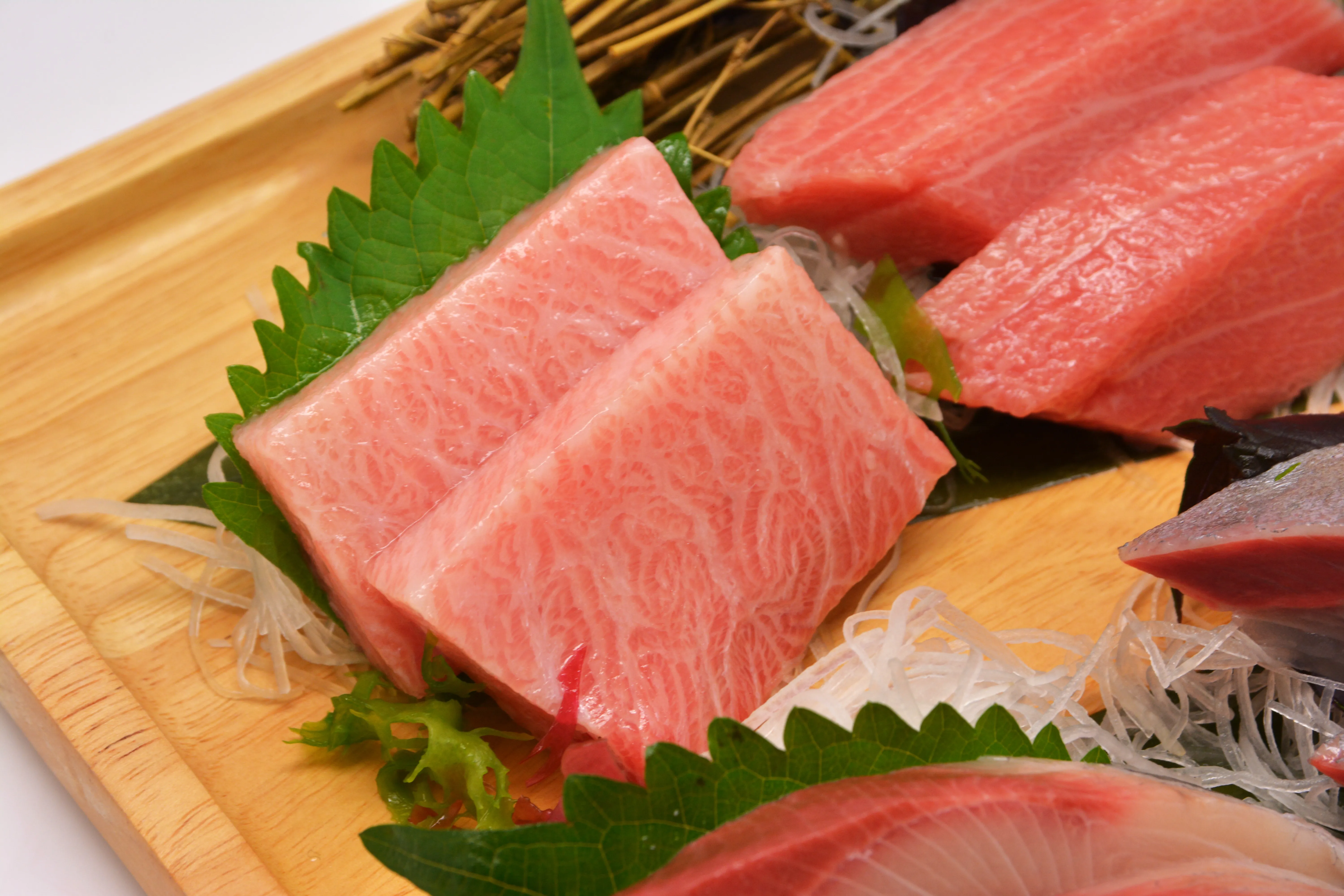 A close-up shot of a premium Japanese sashimi platter featuring expertly sliced otoro and akami tuna, garnished with fresh green shiso leaves and delicate shredded daikon on a wooden serving board.