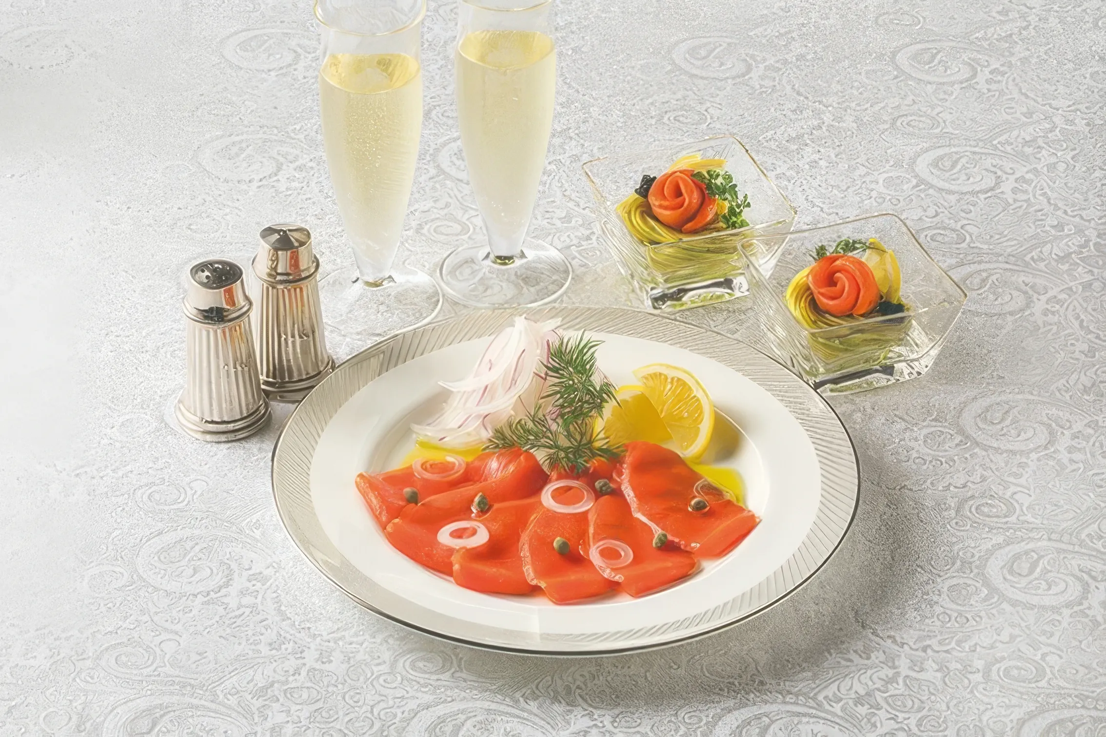 An elegant array of smoked salmon carpaccio slices, garnished with red onion, capers, and dill, alongside two champagne flutes and small salmon rosette appetizers, presented on a white patterned tablecloth.