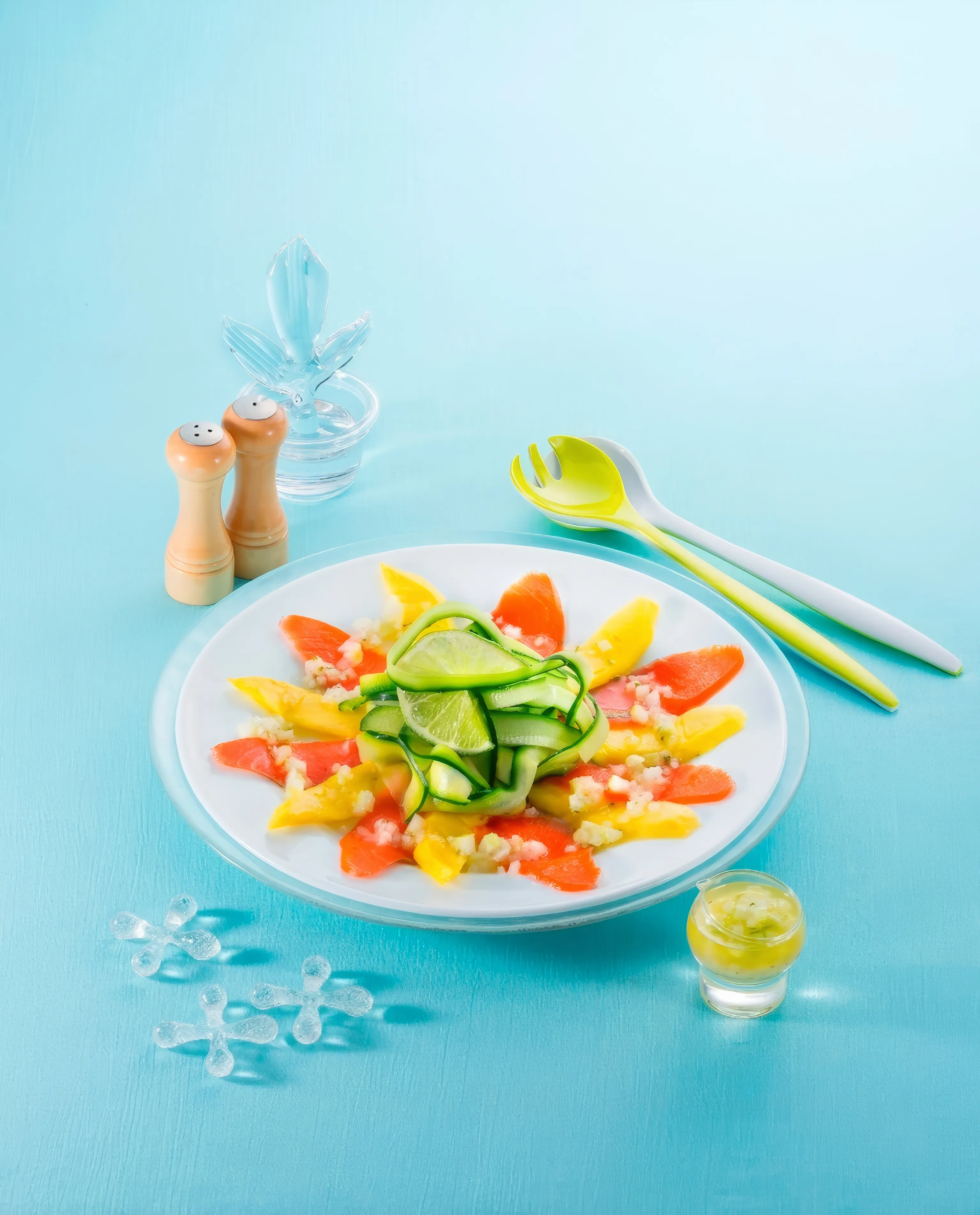 A vibrant smoked salmon and mango salad with cucumber ribbons and lime wedges, served on a white plate against a light blue background. Wooden salt and pepper shakers and salad servers are visible nearby.