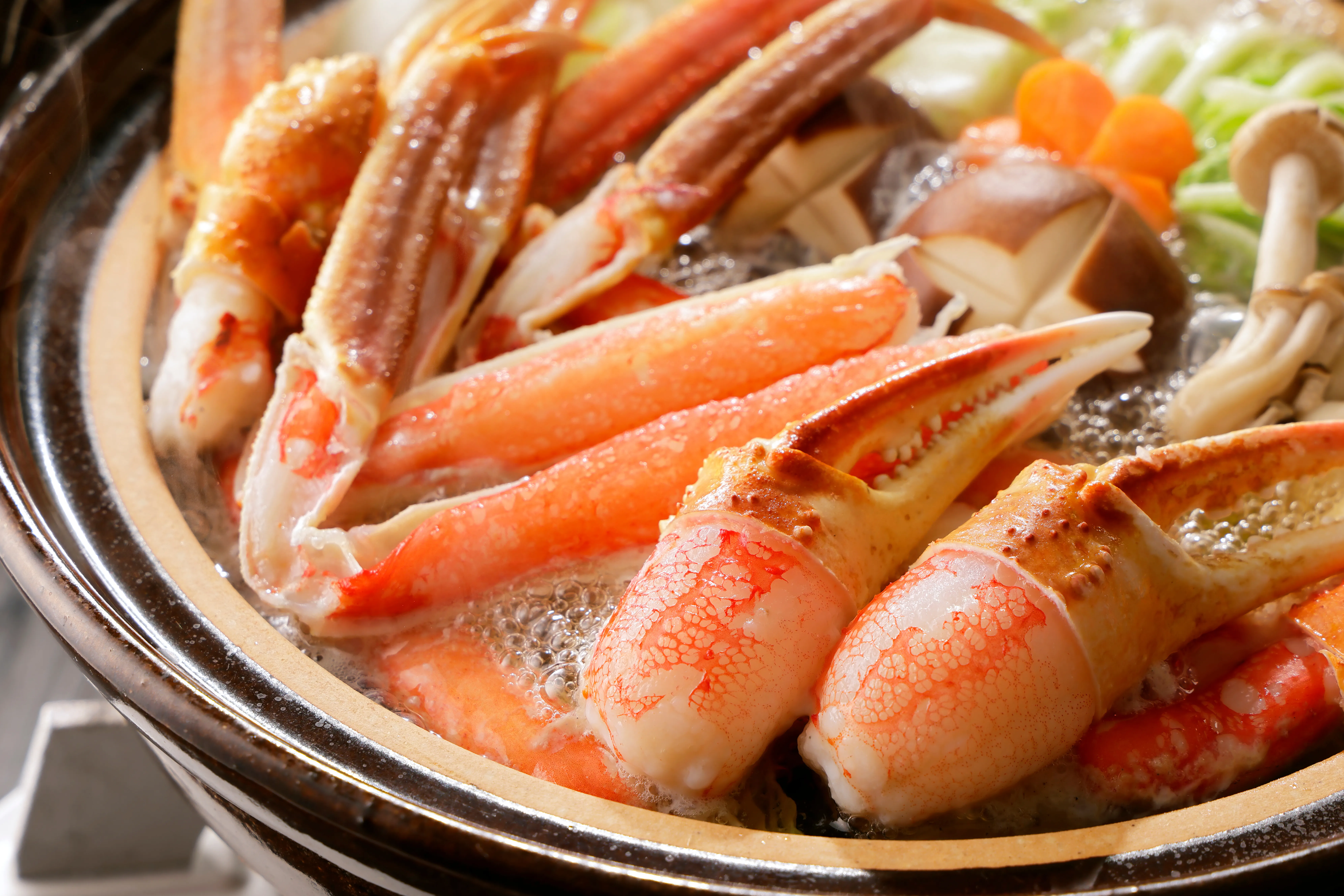 A vibrant close-up of a steaming Japanese snow crab hotpot, showcasing plump orange crab legs and claws nestled among fresh green vegetables, sliced carrots, and various mushrooms in a traditional dark earthenware pot.