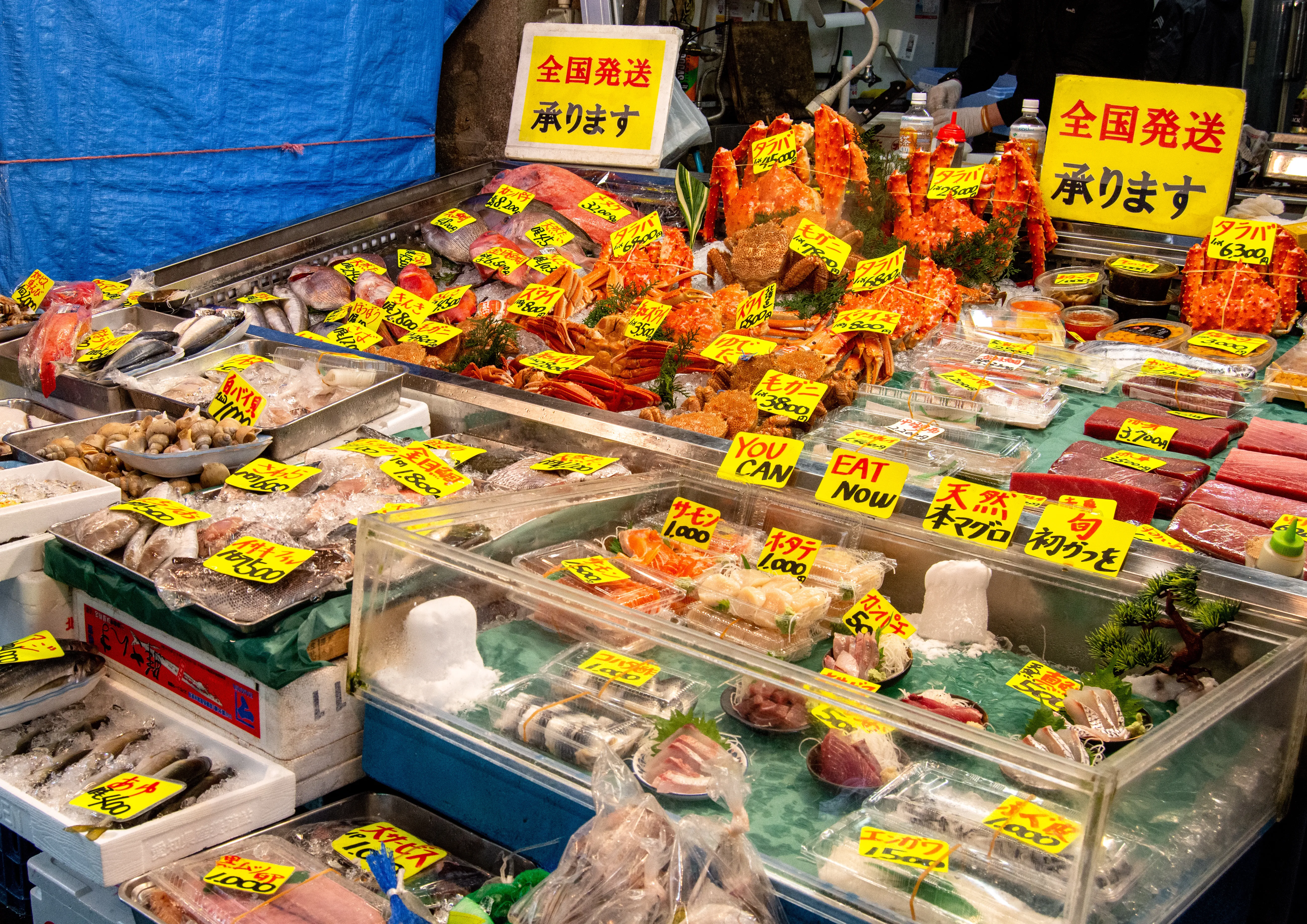 A bustling Japanese seafood market stall showcases an abundant array of fresh fish, crab, and shellfish laid out on ice, with yellow price tags and Japanese signage. Various cuts of tuna and salmon sashimi are also visible in clear display cases, emphasizing freshness and variety.