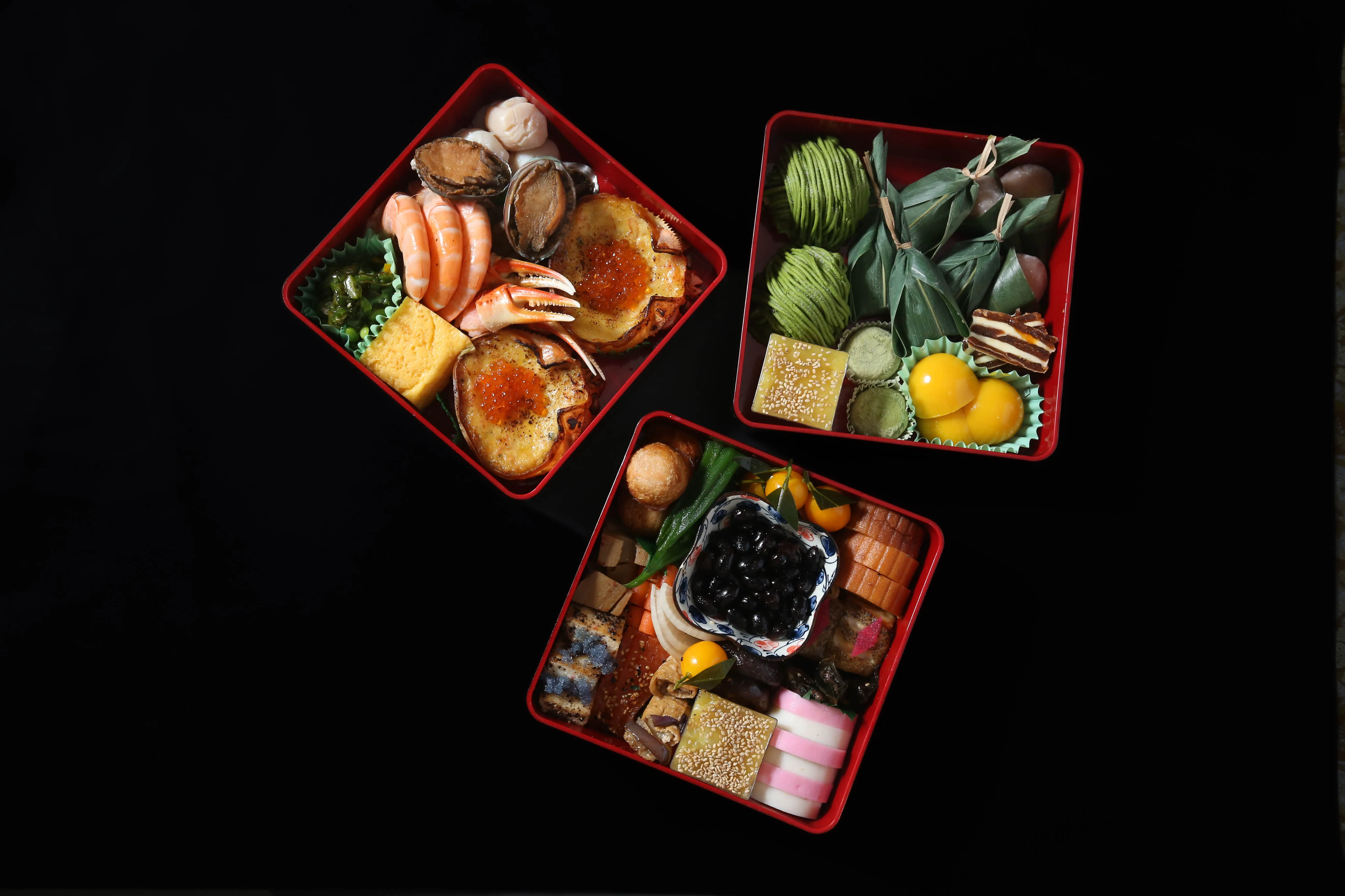 Overhead shot of three red osechi boxes filled with traditional Japanese New Year food on a black background.